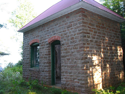 A square brownstone building with a hipped metal roof has an arched door and arched window on one side.