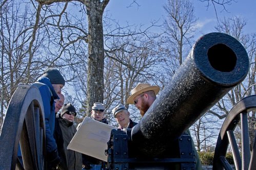 A park ranger and a small group of people gather around an old black cannon. 