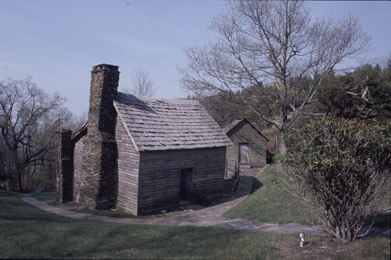 A path leads around a wooden cabin with a single door and a stone chimney. 