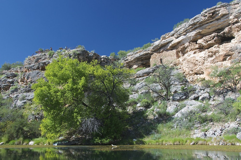 A view of two small, single-story cliff dwellings from the water of Montezuma Well. The banks are surrounded by grasses, cattails, rushes, and a few trees.