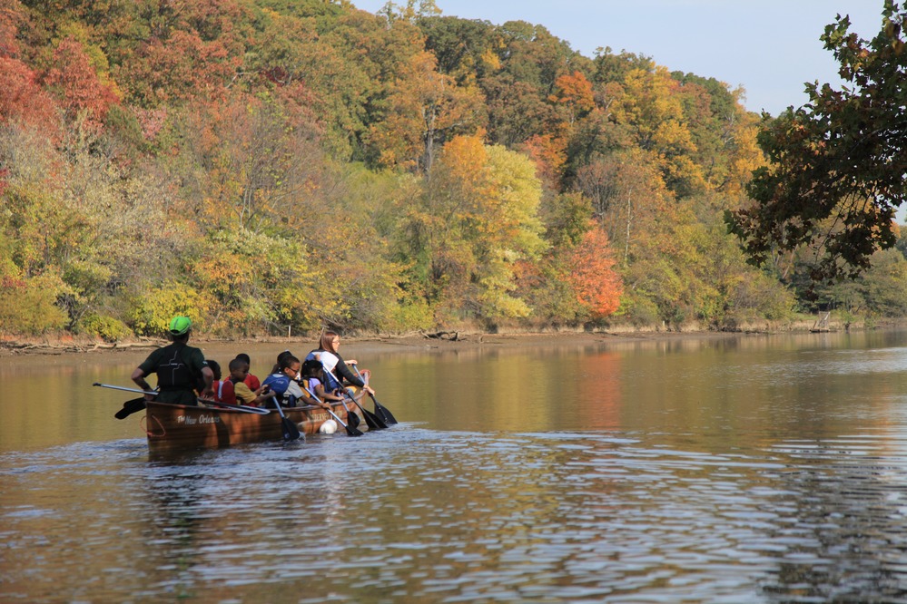 Children and chaperones row a canoe up a river past trees with fall colors