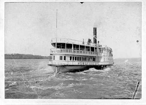 Boat on the river.  Has two decks, the lower deck has windows around it and the upper deck is open.  A smoke stack in near the front of the boat.  