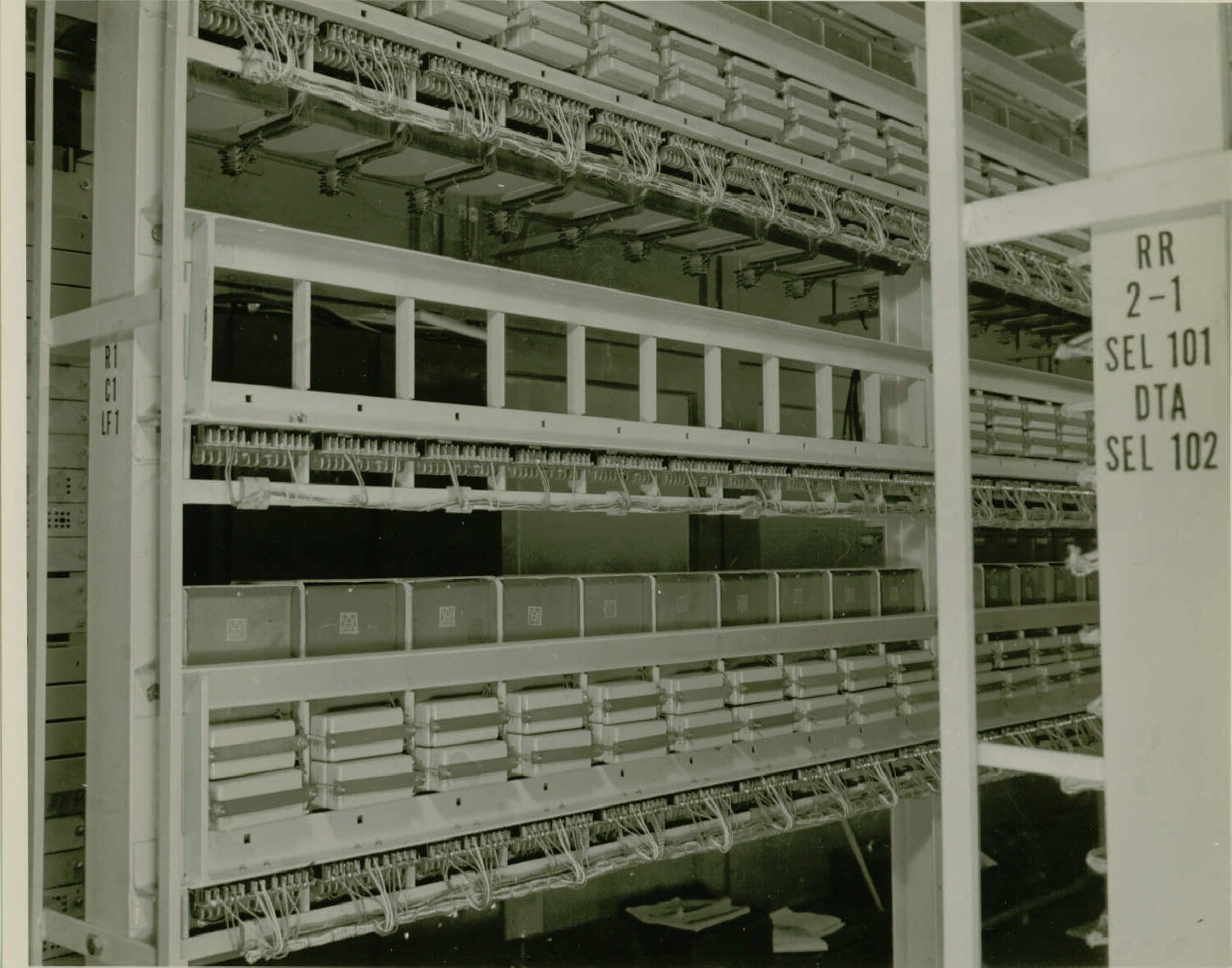 Black and white photo of a metal rack. The rack has three rows. The rows have spaces for boxes to be mounted. Each space has wires that can connect to the box and then runs along the bottom of the row towards the back of the rack. The middle row has 10 spaces with no boxes mounted.