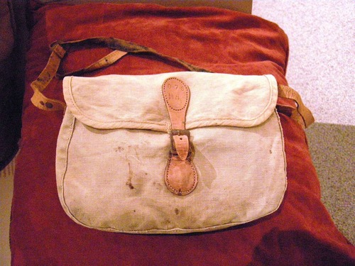 A fisherman's creel bag, which is light pink in color and has a leather strap and metal buckle.
