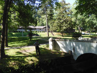 In the foreground, a white stone bridge with someone looking into the water. In the background, the stone maintenance building surrounded by trees.