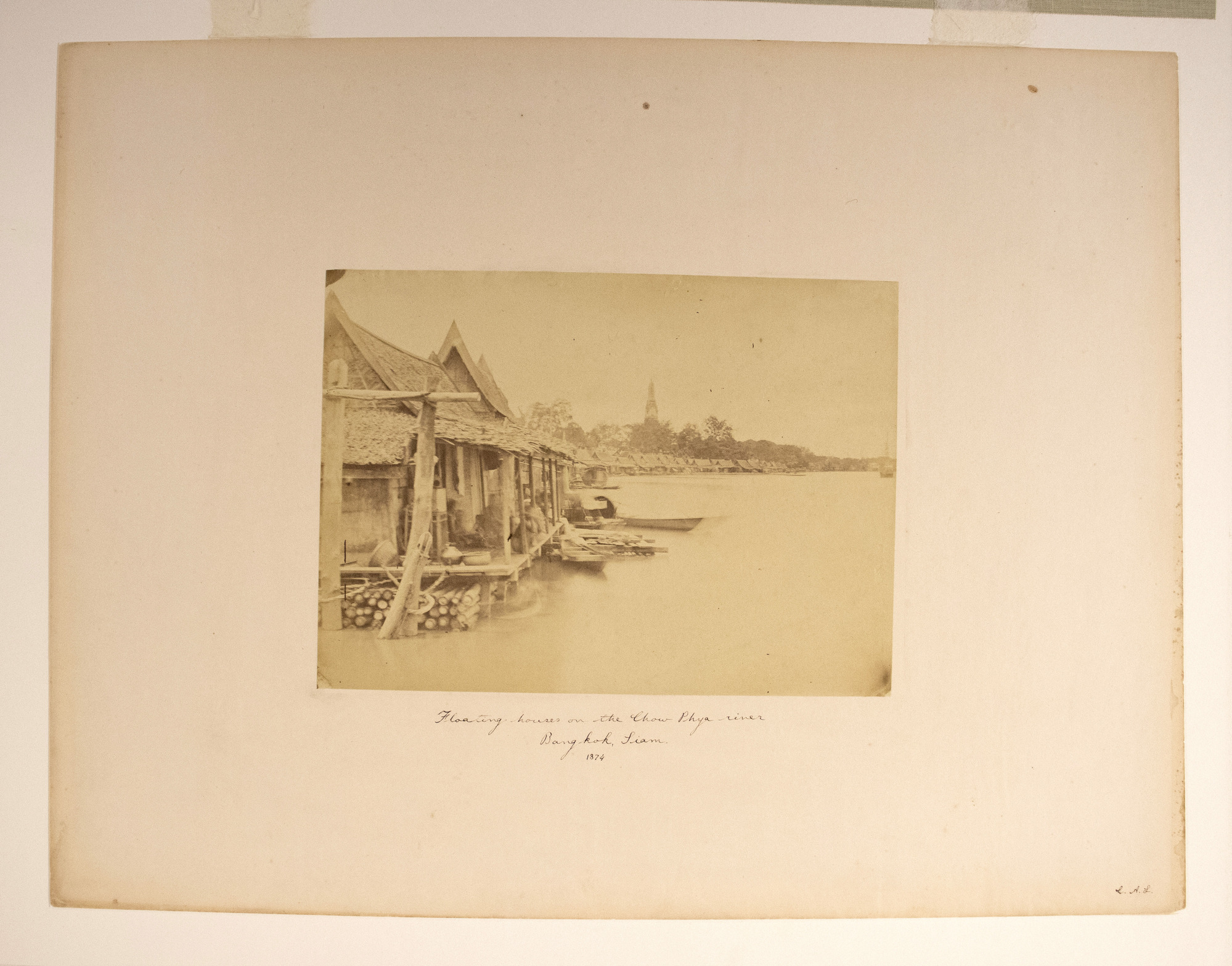 Sepia photograph with an inscription representing houses on water, boats, a few people and a row of houses and trees in the background.