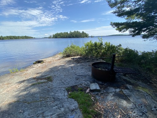 View of houseboat site core and of the fire ring with a couple of branches next to it overlooking the water. A couple of tree covered islands are in the background across the water.