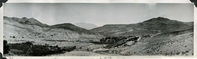 This is an historic black and white panoramic photograph from the Scotty's Castle Historic Photograph Collection, Death Valley National Park of vegetation in the left foreground, tent structures on left background, fencing, a large two-wing building under construction, landscaping, and other structures in a desert landscape. Black ink inscriptions along lower border.