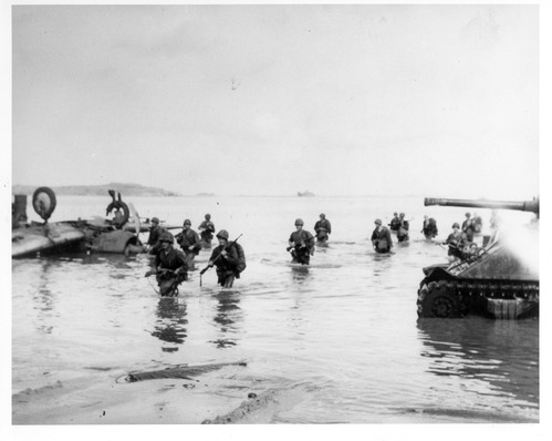 A line of male soldiers in combat gear and holding guns walk through waist high water. On their right is a tank and on their left is an upturned plane.
