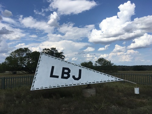 Under a sky of puffy, white clouds, a gray triangular pyramid sits with dark letters L-B-J on its side. in a fenced area.
