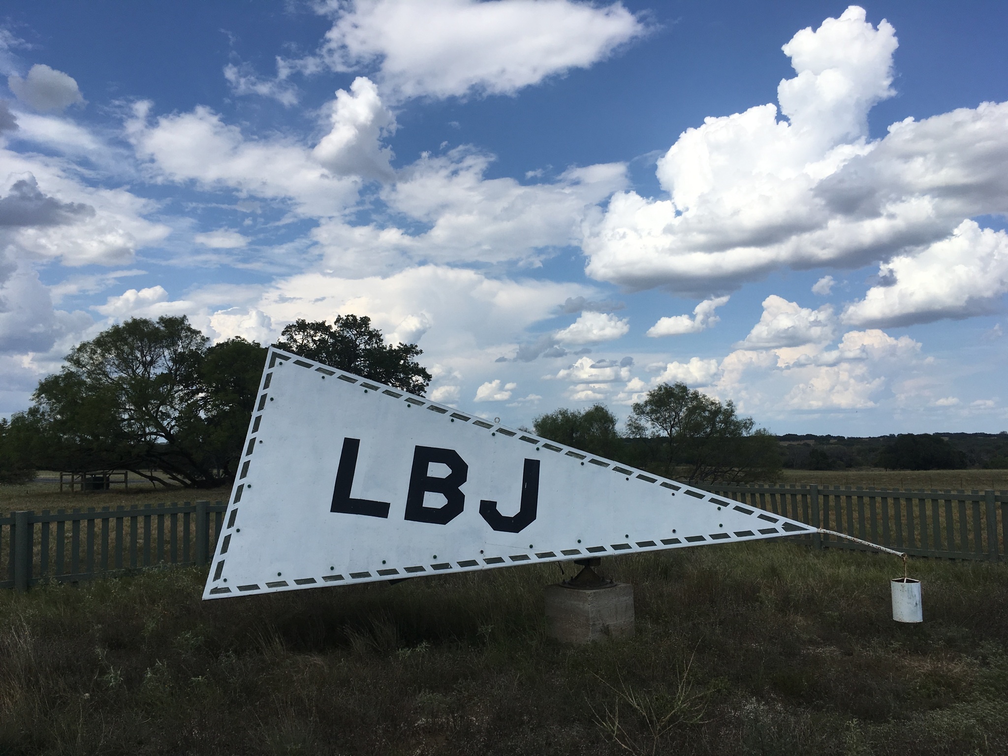 Under a sky of puffy, white clouds, a gray triangular pyramid sits with dark letters L-B-J on its side. in a fenced area.