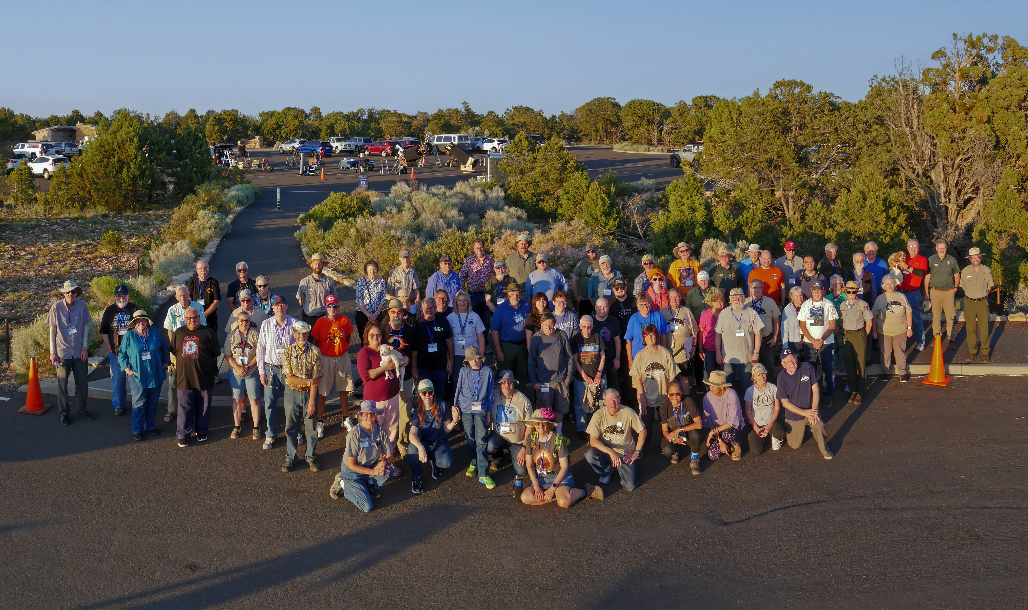 Just before sunset. a group of around 70 people are posing for a group photo on the edge of a large parking lot adjoining a forested area. 