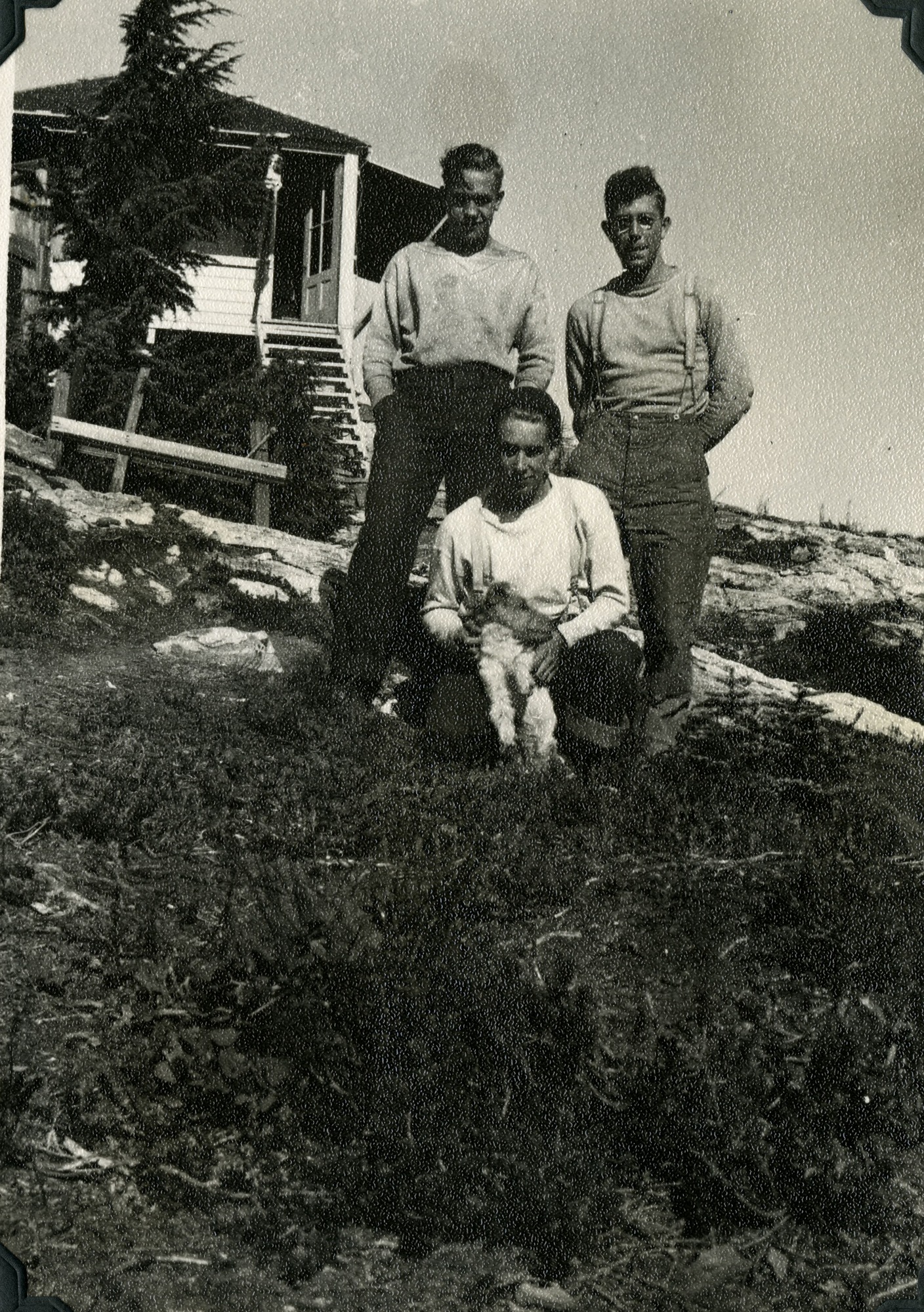 Three white men and a terrier standing in rocky terrain.  A wooden structure is in the background.