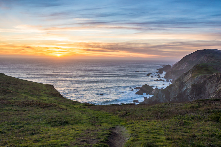 Wide view of the sun setting over the ocean, framed by grassland and cliffs in the foreground.