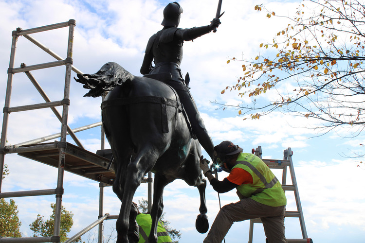 Two men stand on a ladder and scaffolding platform to perform maintenance on the Joan of Arc statue.