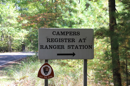 A rectangular tan sign reads “Campers Register at Ranger Station.”  A black arrow points to the right.  On the sign post is a small brown triangular sign with the National Park Service arrowhead and the words “Fee Area.”  Green tree branches fill the background.  