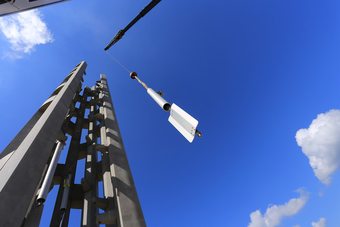 Deep blue sky offsets a dangling metal chime tube with waving metal sail at the bottom. Chime is moving toward gray tower with many windows.