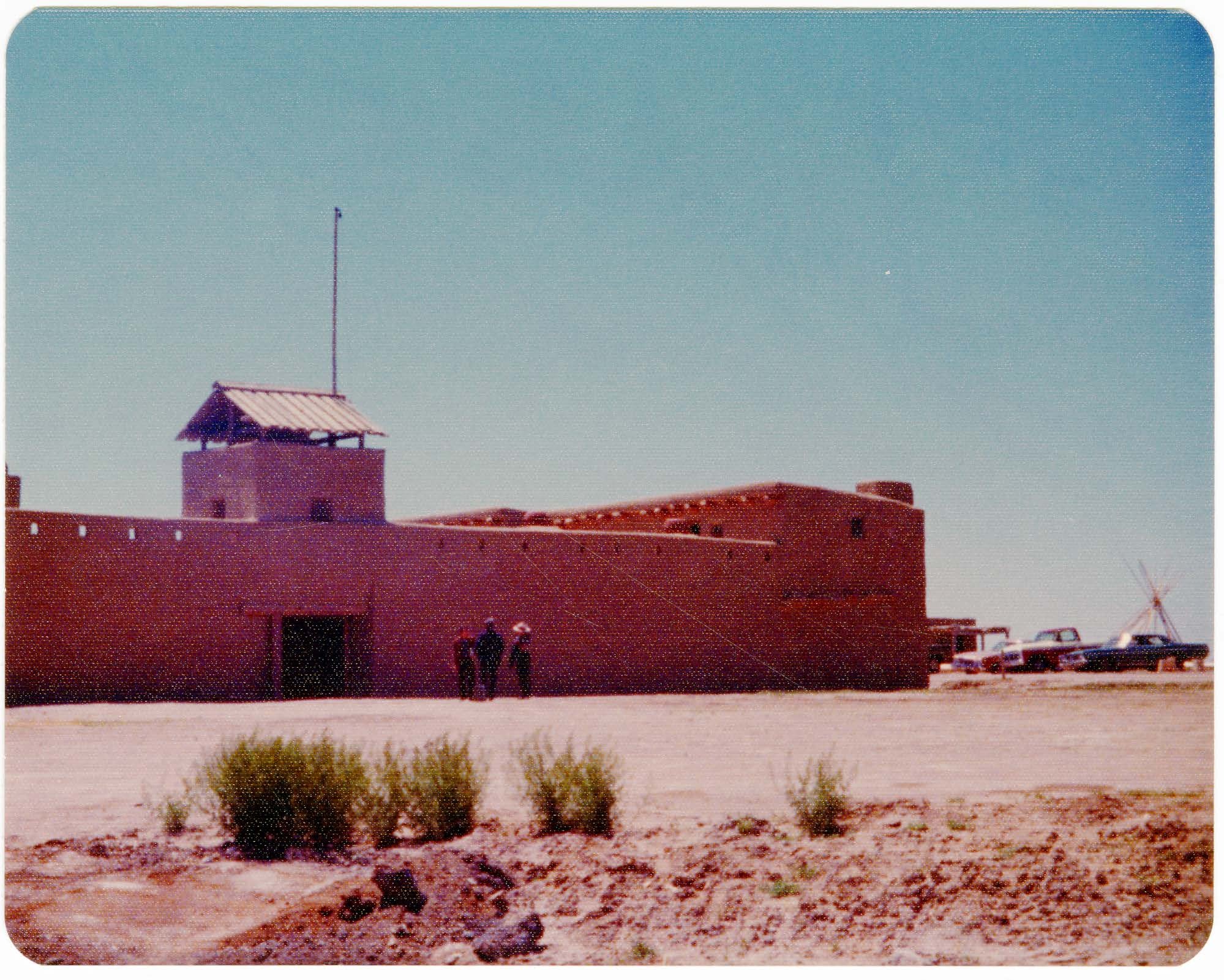 Taken as construction was nearly completed, this image shows a group of people standing near the front gate of the fort. To the right side of the fort a circa 1965 adobe blacksmith shop is visible behind the reconstructed fort along with a number of automobiles. 