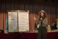 A park ranger gesticulates while standing in front of a board with a map and an outlined schedule of events for a cruise ship day in Glacier Bay.