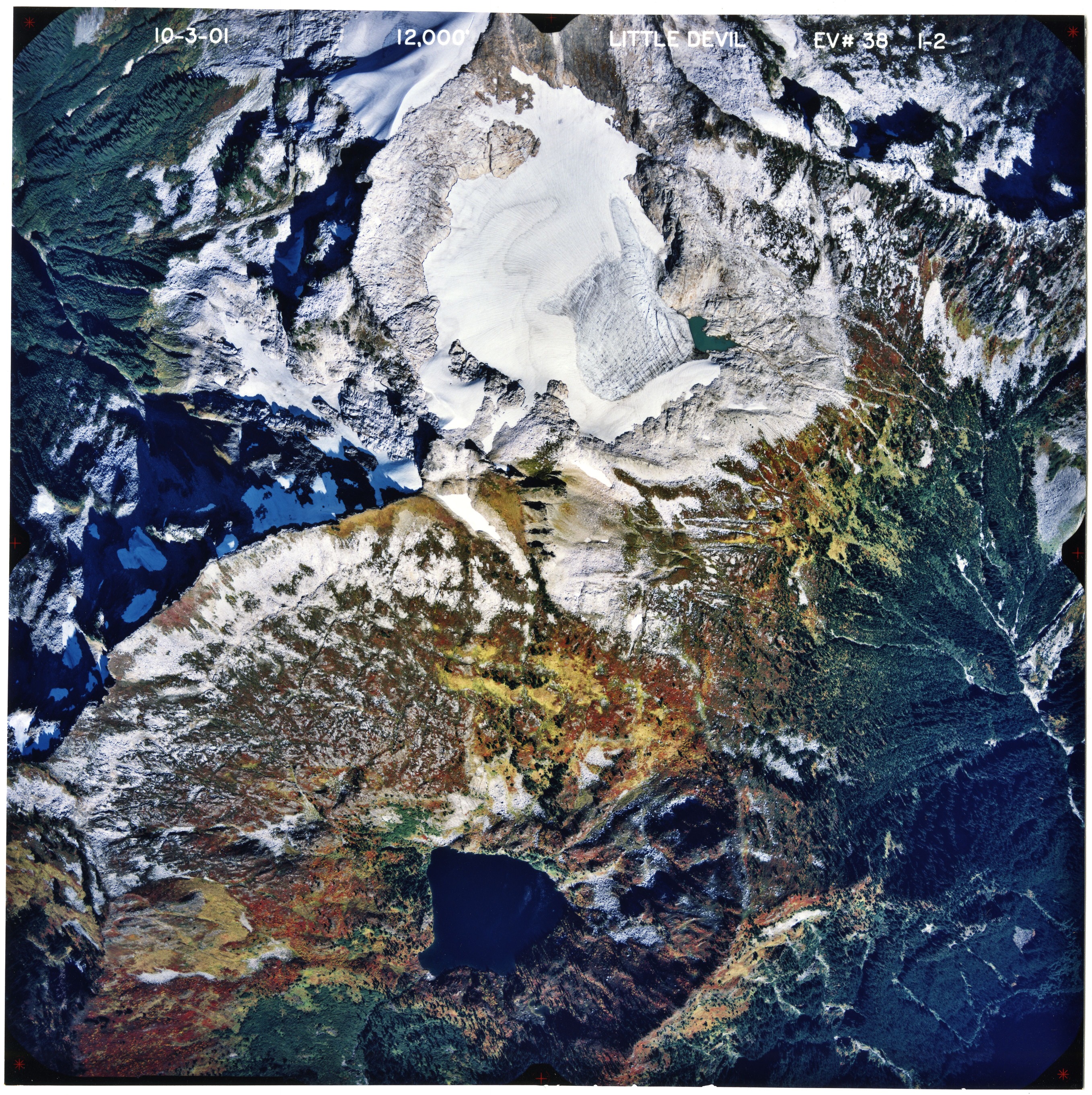 Color aerial photograph of glaciated mountain peak with alpine lake, forested valleys below.