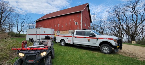 An ATV, utility truck and cargo trailer sit in front of a red farm barn