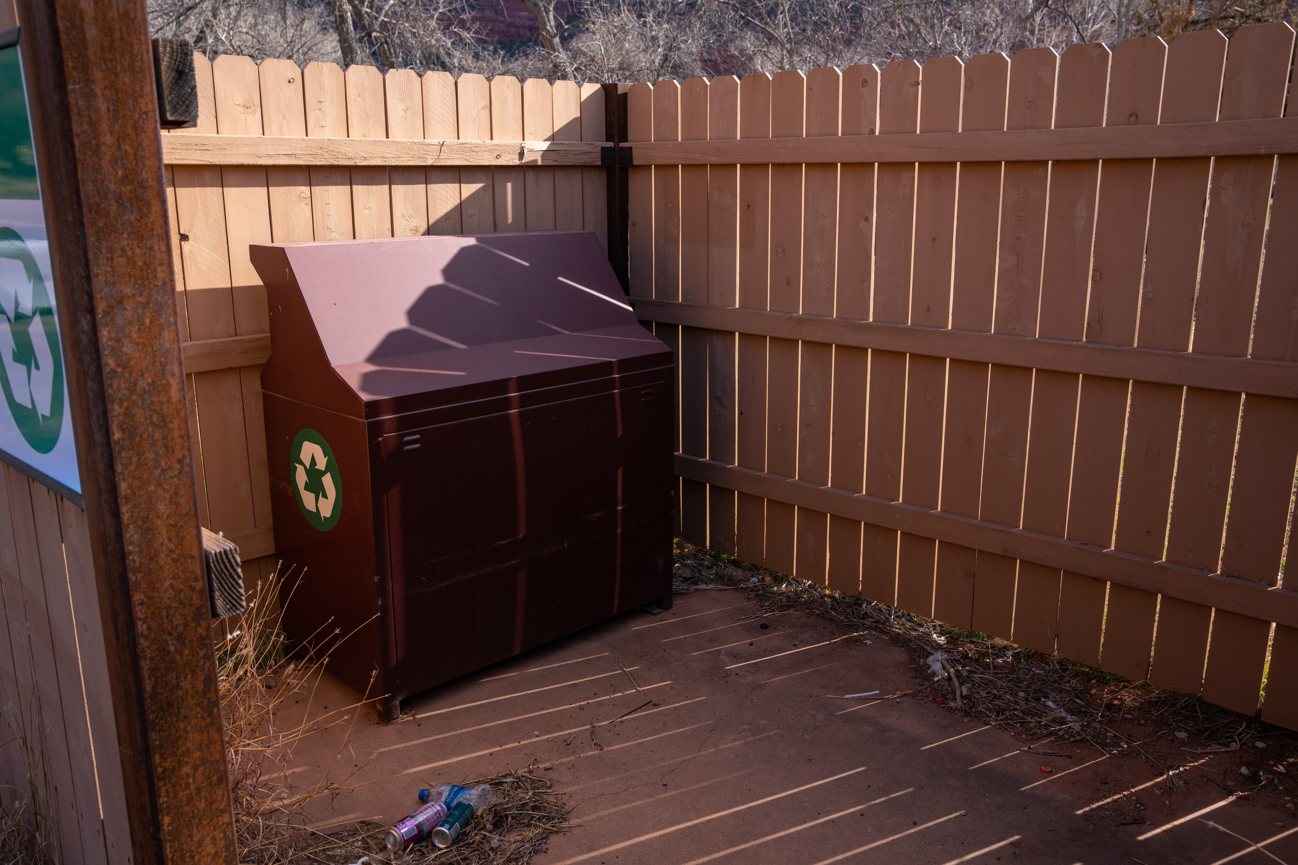 A fenced in brown trash can with a green recycling symbol on the side surrounded by weeds and trash