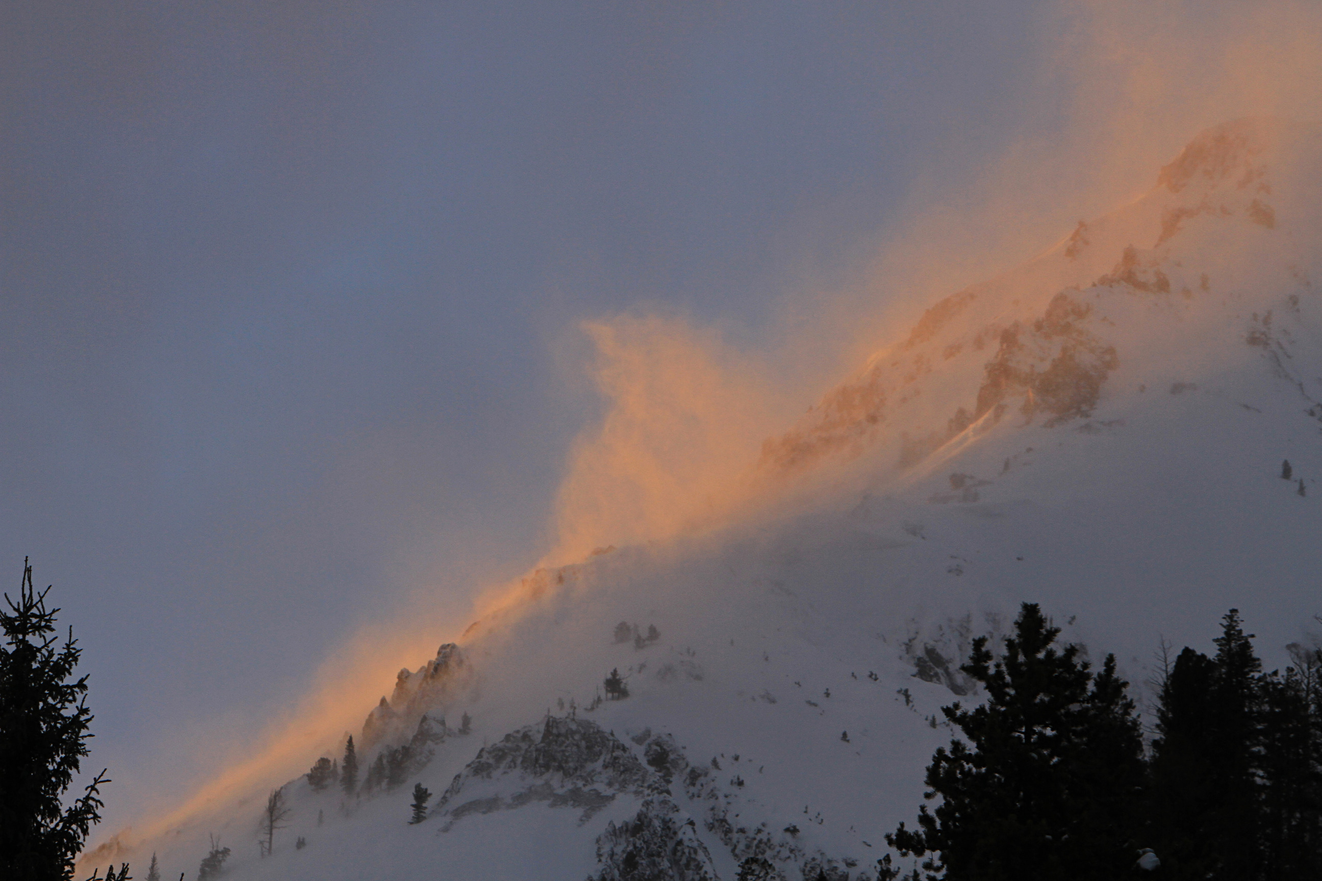 Close up of the side of a mountain while snow is blowing off of it.