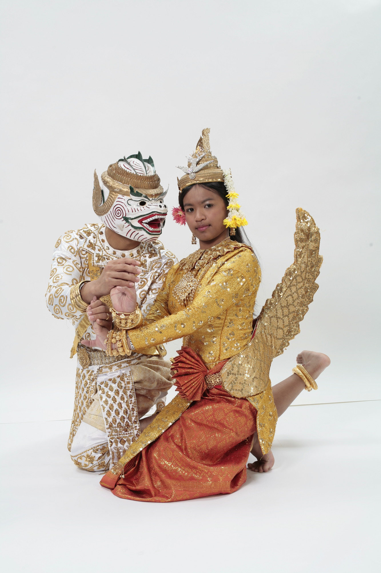 A dancer claps the hands of another as they kneel down. Hanuman's dancer wears a white monkey mask with a golden head ornament. His face has red spiral markings, and his smile and eyebrows are highlighted in green. His outfit is an elaborate embellishment of gold patterns. The Maccha dancer wears a golden crown which spirals upward. Her tunic is gold with golden embellishments, while her skirt is orange. She wears a golden mermaid tail tied to her waist. 