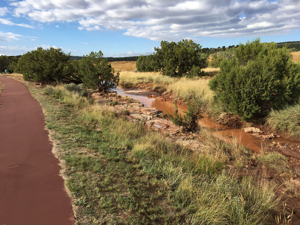 A red asphalt trail passes by a creek bed containing opaque, muddy, reddish-brown water.