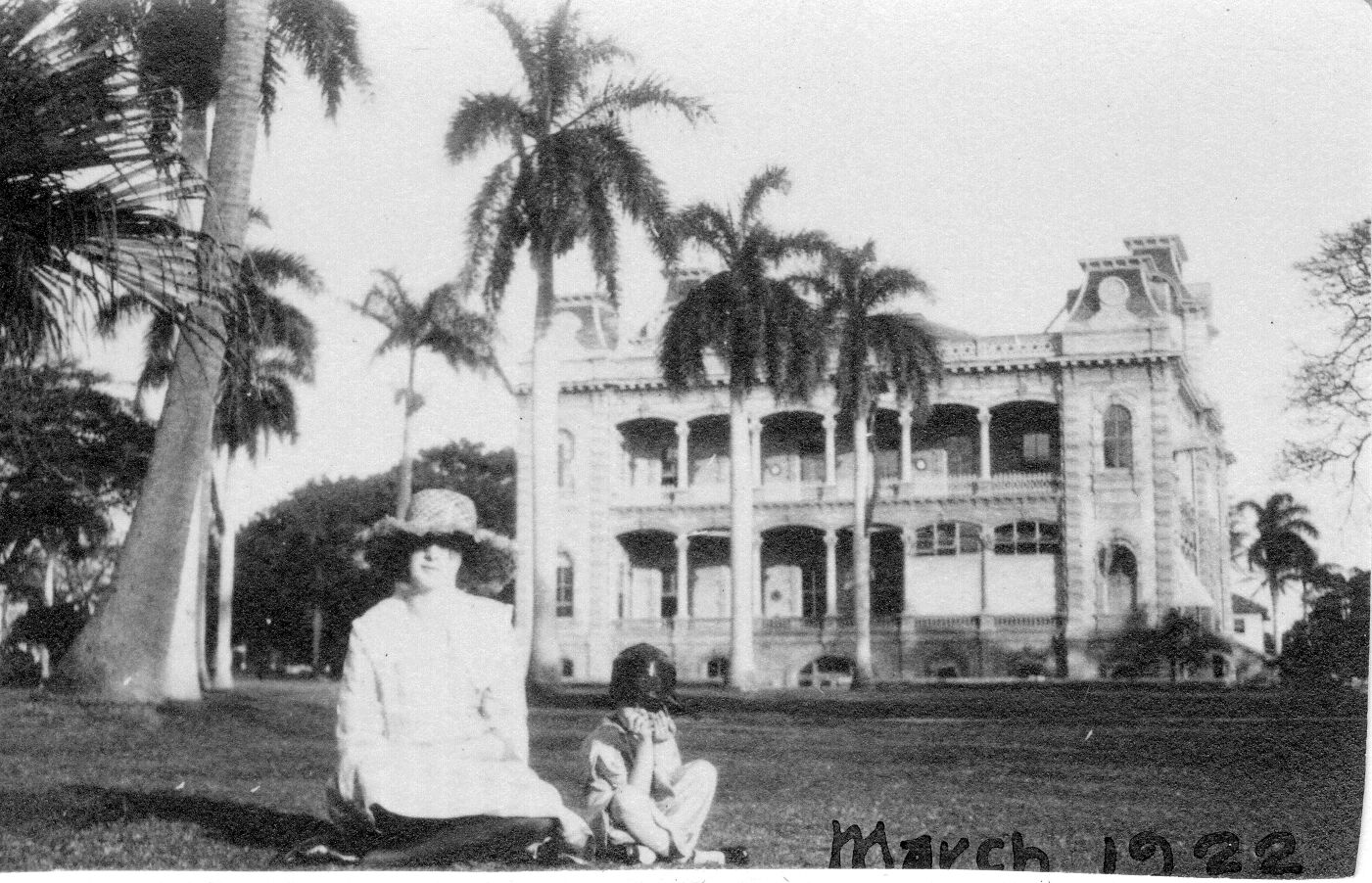 A black and white image of a woman and girl sitting on the front lawn of ʻIolani Palace. The woman and child are pictured in the foreground of the image. The woman is wearing a wide brimmed hat and a long-sleeved dress. She is looking towards the camera. To the left of her is the girl. She is wearing a hat and a short-sleeved dress. Her knees are raised slightly with her arms in front of her face pulling her hat over her eyes. In the background there are palm trees and the front of ʻIolani Palace. The palace is a two-story structure in the American Florentine style. It is built out of brick with four corner towers and two center towers with a lanai on both levels. Along the bottom edge of the photo, "March 1922" is written in ink.
