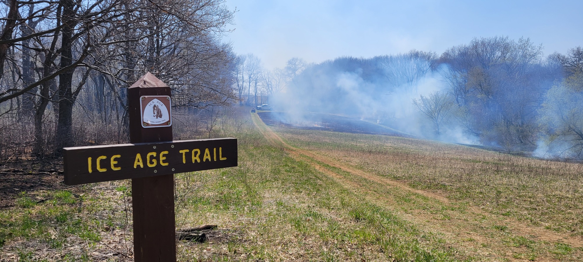 Wooden Ice Age Trail sign with burned ground and smoke in the back ground.