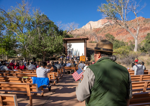 Photo of a man in a green vest and a tan flat-hat standing in front of an amphitheater full of people. A stage is seen in the distance, under a clear blue sky. 