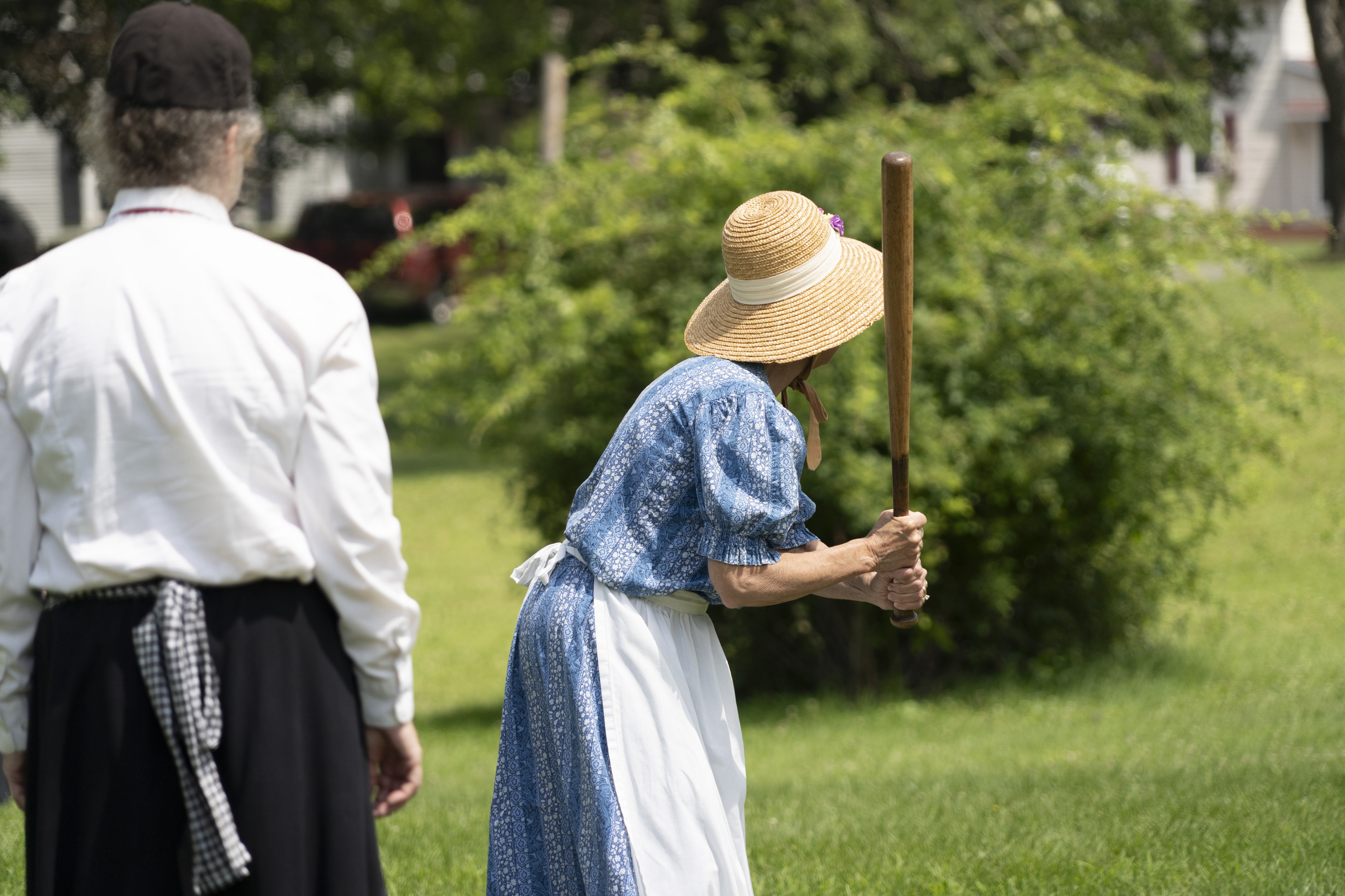 A woman in a 19th century baseball outfit prepares to swing a baseball bat.