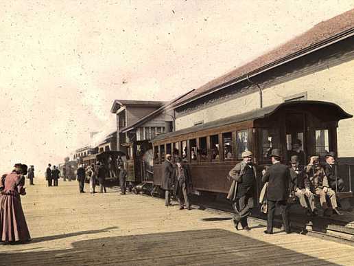 Woman in a red dress nears a train at a station. Three men dangle their legs off the caboose. Others wander the platform.