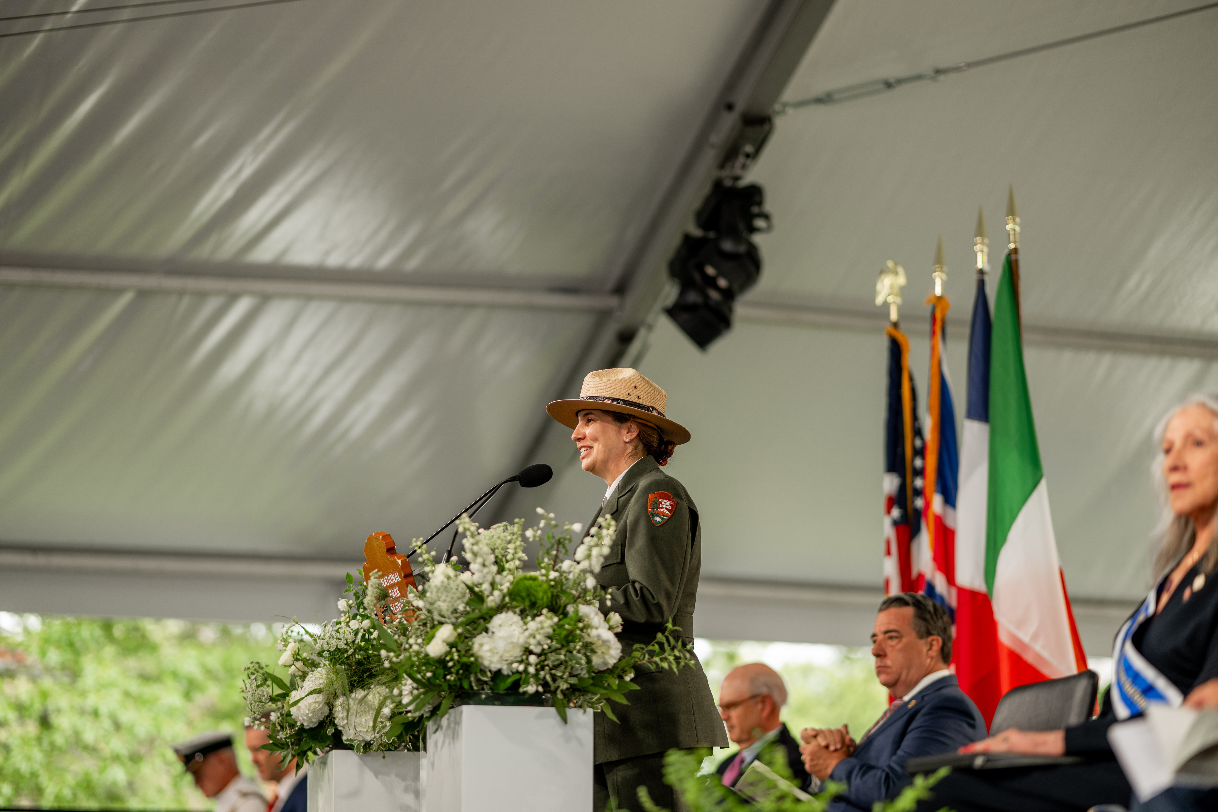 A National Park Service Superintendent in dress uniform speaks at a clear podium. A National Park Service arrowhead hangs from the front of the podium. Flowers are on either side of the podium.