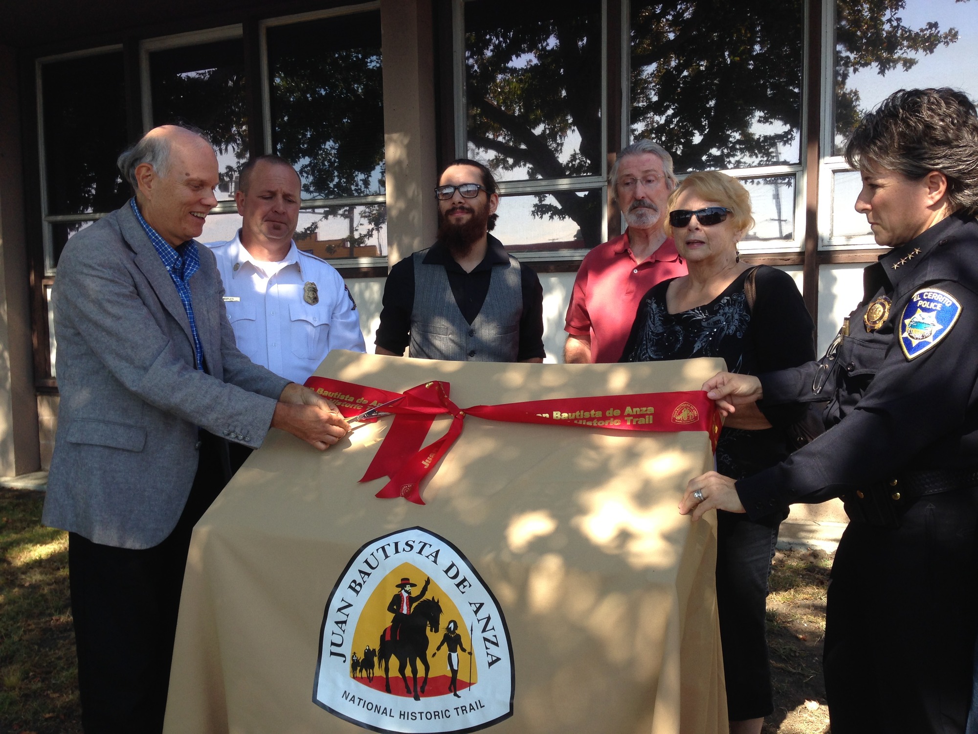 A group of people stand next to a sign covered in a brown cloth with the Anza Trail insignia on it while one person smiles and cuts a red ribbon tied around the cloth