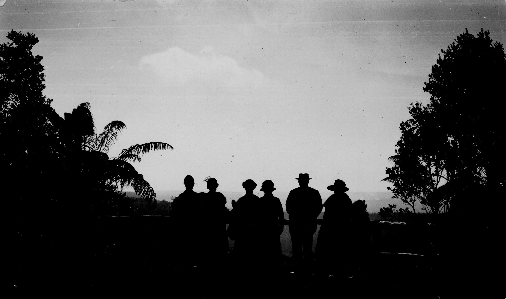 Black and white photograph of eight people posing for the camera by a lookouti. The photograph is very dark. The crowd is standing in a clearing with their backs against a wooden railing. Beyond the railing, Kīlauea crater is very faint, but partially visible. A single cloud is present in the sky and vegetation lines the left and right borders.