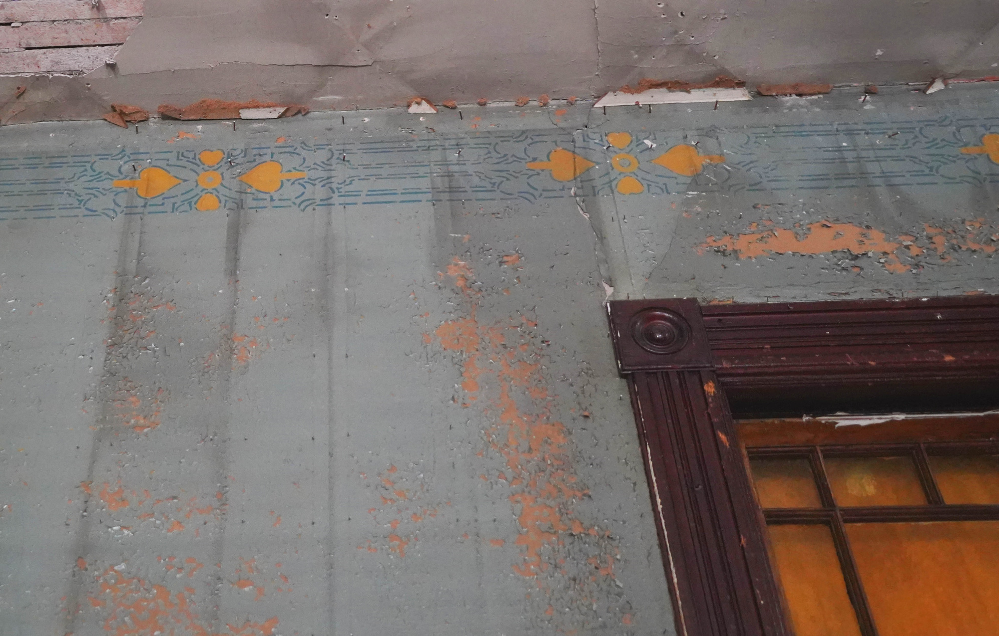 Detail of stenciling at top of north wall.  The wall is a light blue/green with with a darker blue geometric or scroll motif at the top. A bright yellow circle and spade pattern repeats at about 10-inch intervals. The top corner of a window is also visible in the lower right corner of the image.