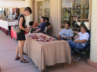 People seated at and standing in front of tables set with various objects in front of windows in the shade.