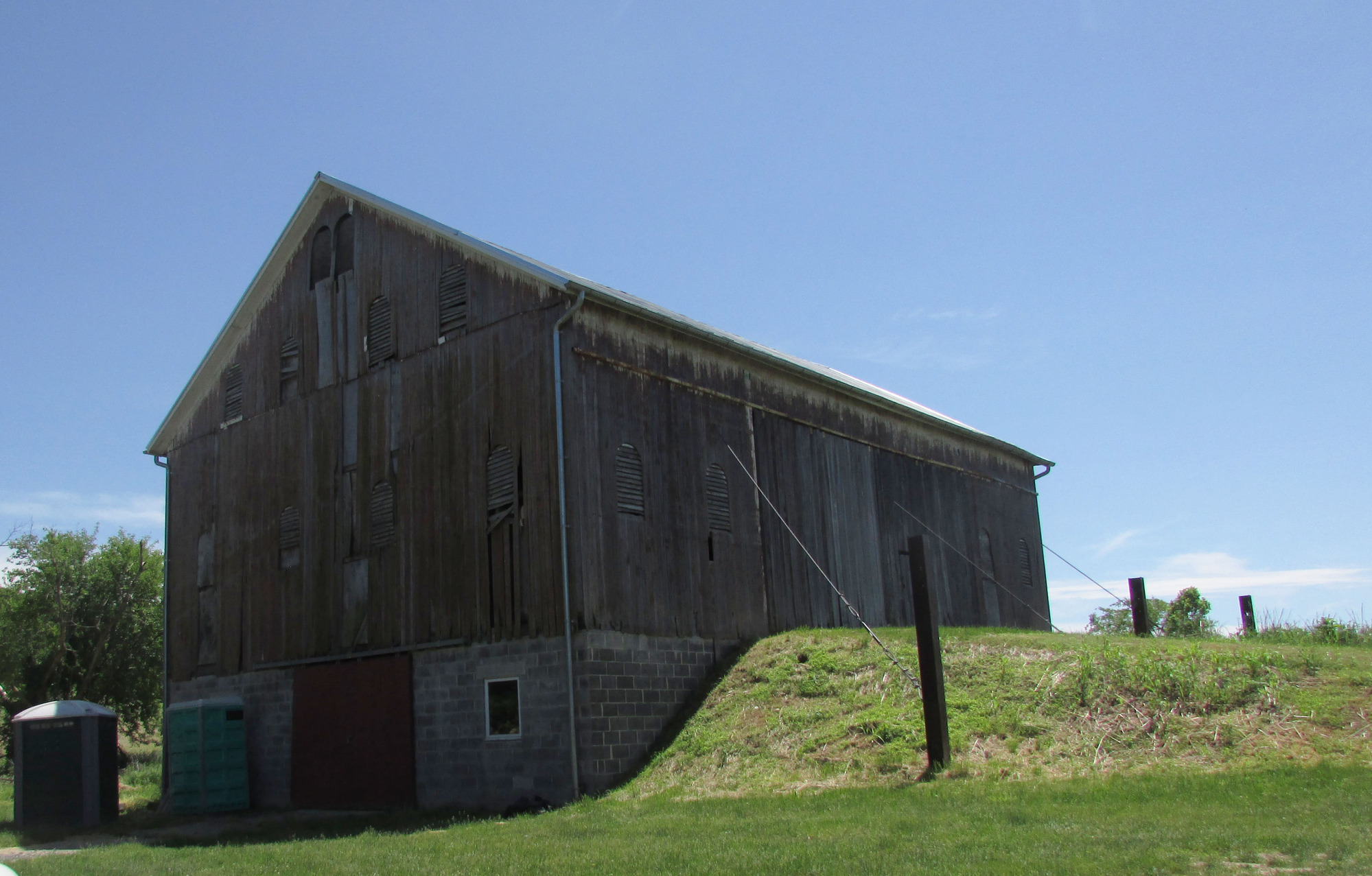 A tall wooden barn stands against the side of a bank of earth. It has vertical board siding, a concrete block foundation, and peaked gable.