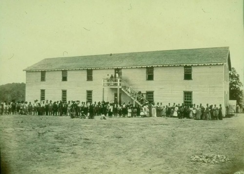 Gray scale photo of wooden school building with a large group of individuals standing outstanding outside of the school building.