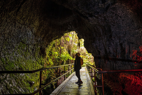 A person standing on a wooden walking bridge inside the entrance of a lava tube. The lava tube is illuminated from outside.
