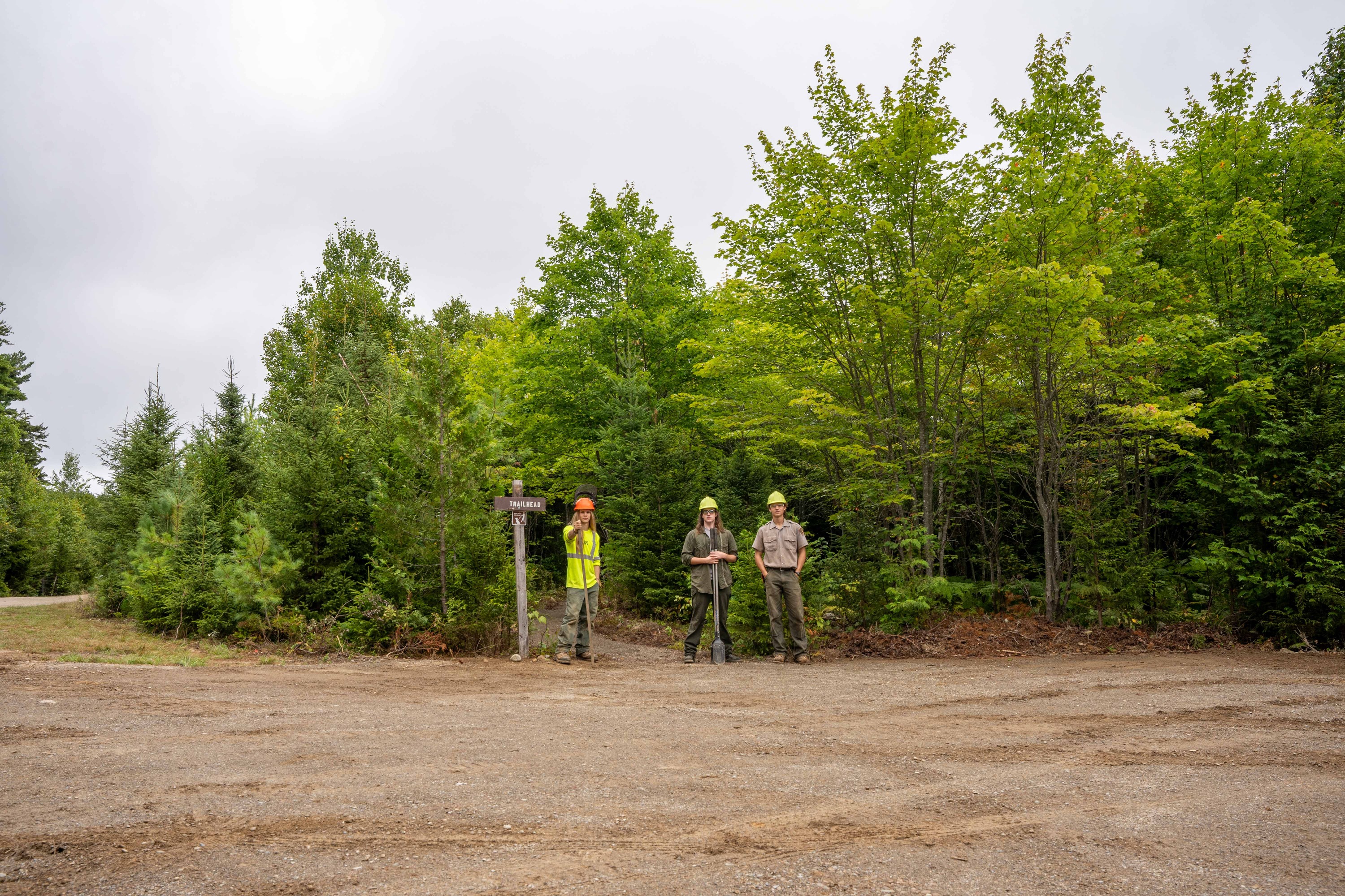 Three trail workers stand at the entrance to Lynx Pond next to the trail they completed. Green trees and cloudy sky fill the background.