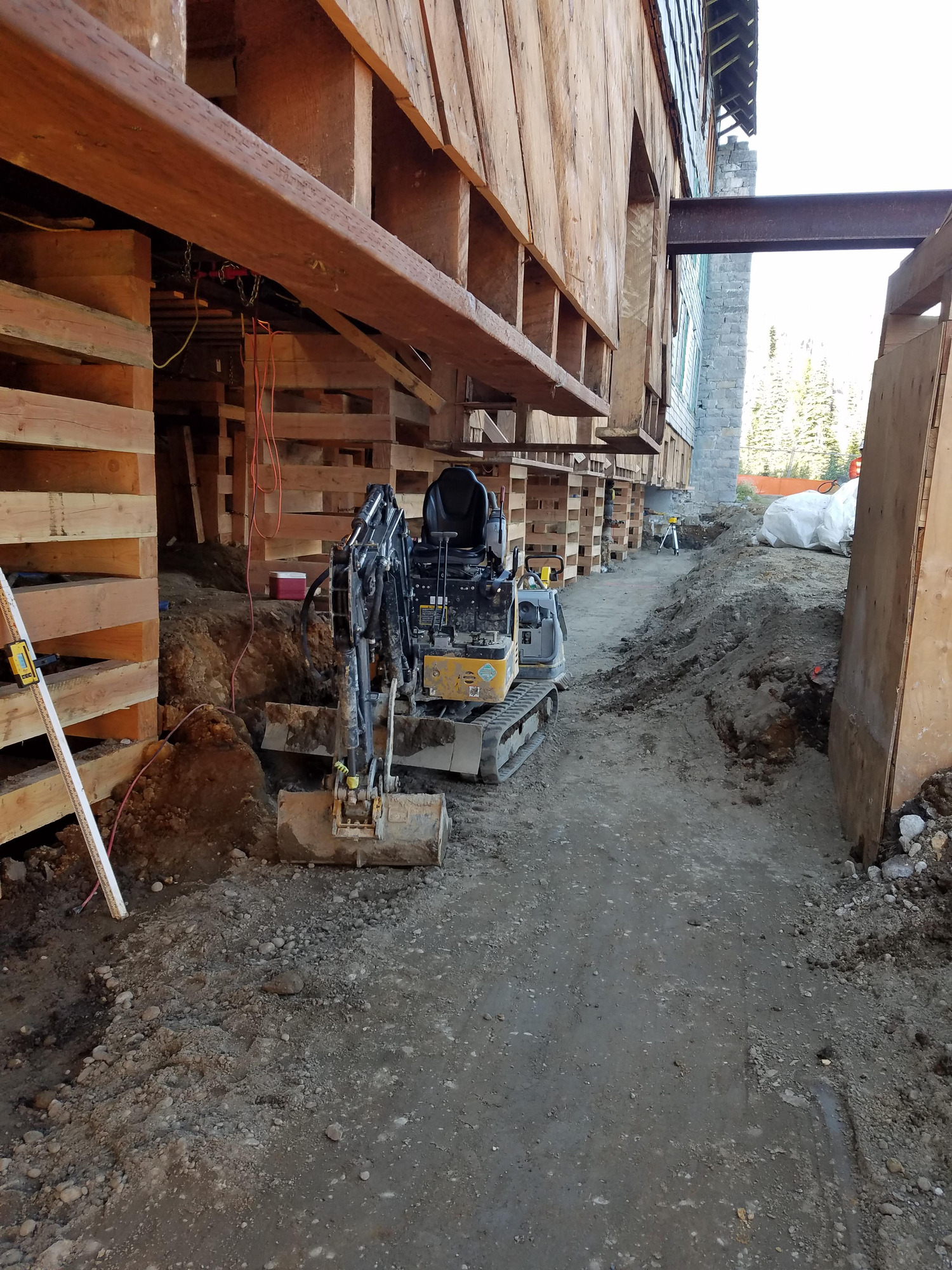 A large wooden building lifted up on wood supports with a tractor digging out the foundation underneath. 