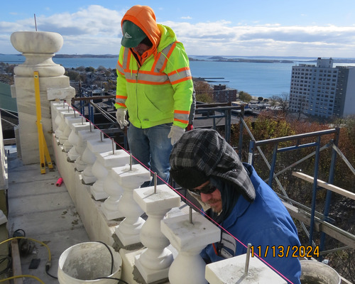 Two workers standing on scaffolding installing short decorative pillars along the side of an upper level of a monument.  