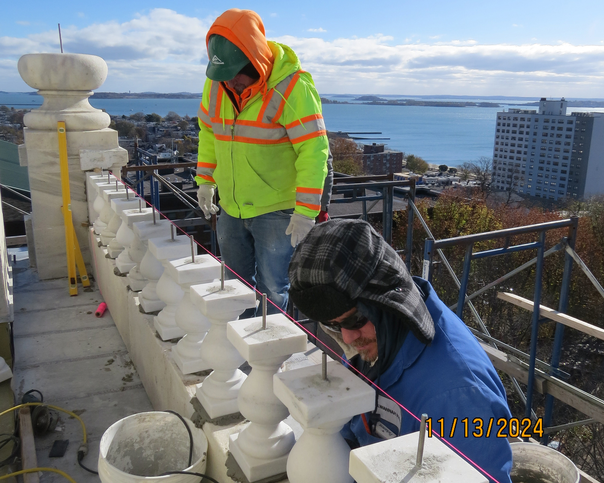 Two workers standing on scaffolding installing short decorative pillars along the side of an upper level of a monument.  