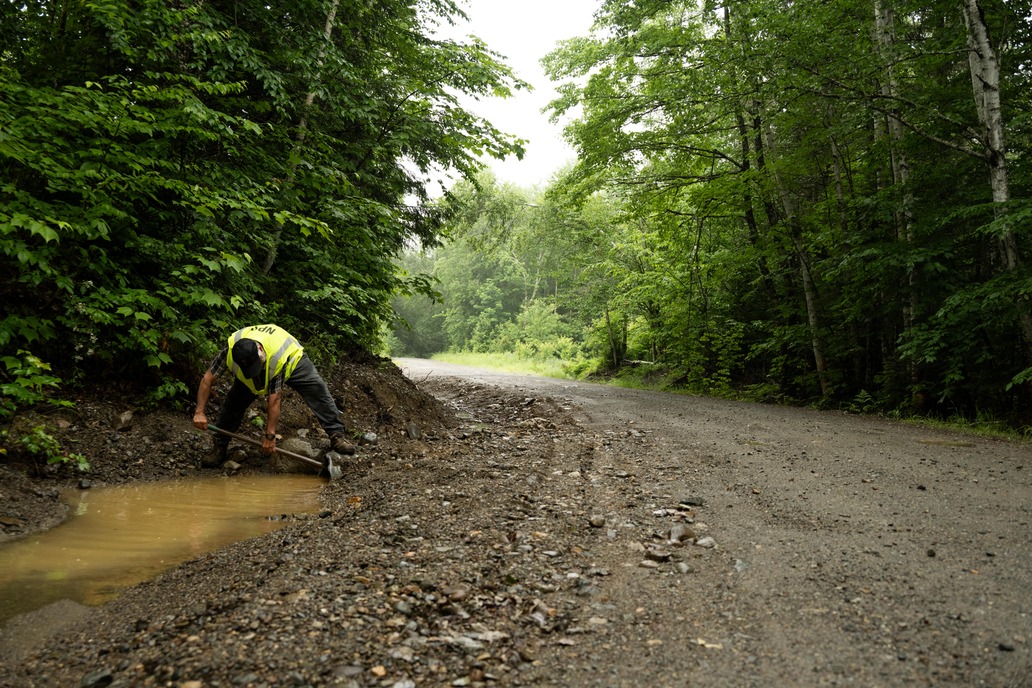 A roads crew worker shovels debris out of a road culvert.