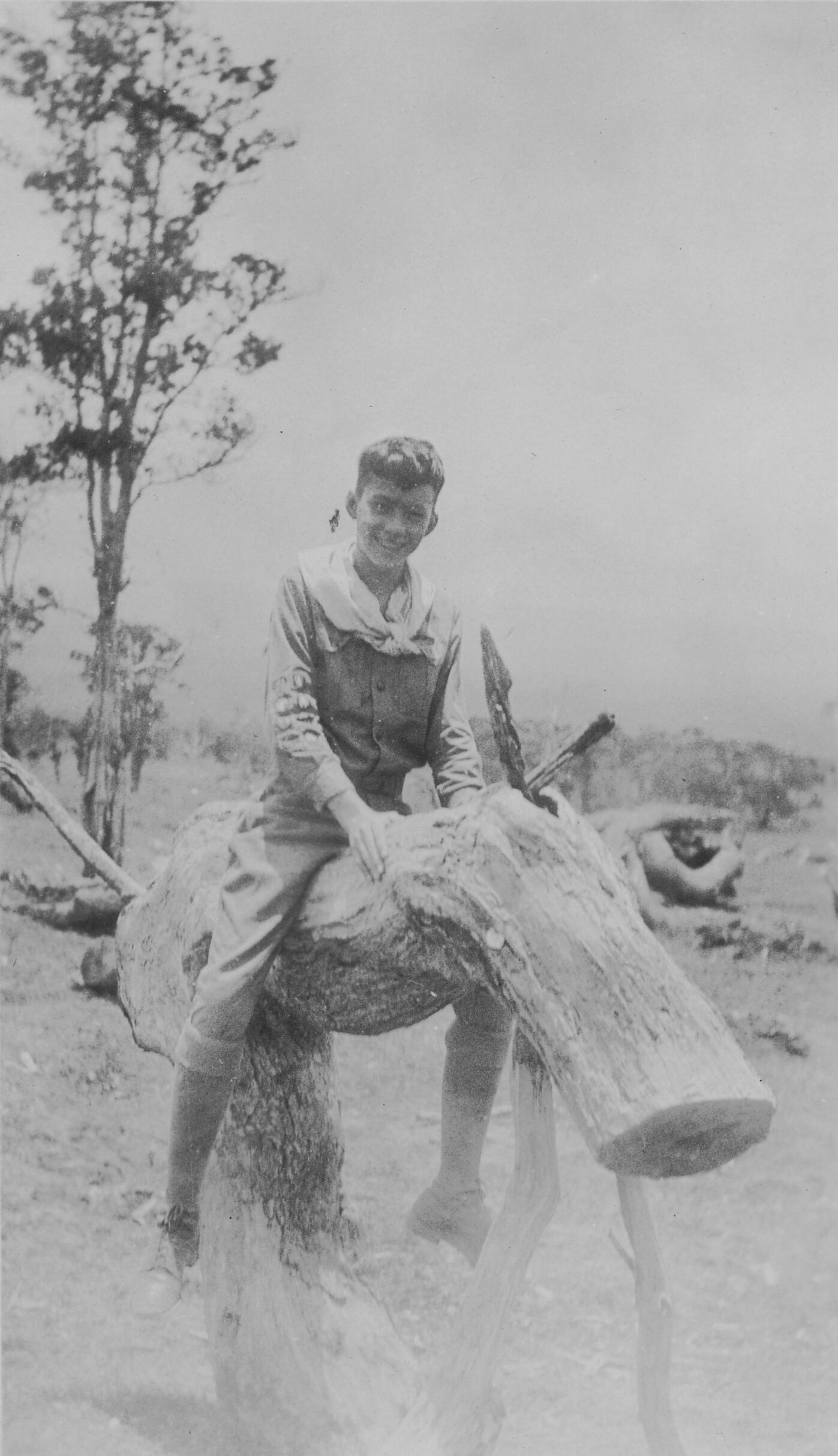 Black and white photograph of a young boy, identified as John B. Moffett, sitting on a curved trunk formation that resembles a horse nicknamed “Spark Plug.” Moffett is mounted on the trunk as if to ride a horse, smiling at the camera. A sparse plane is visible in the background with a singular tree to the left of the image.