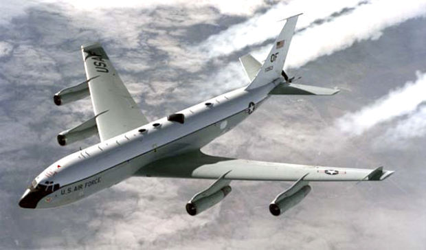 Large plane painted pale green gray and white with the words US Air Force along the side and Air Force markings on the wings and sides, flying through the air.