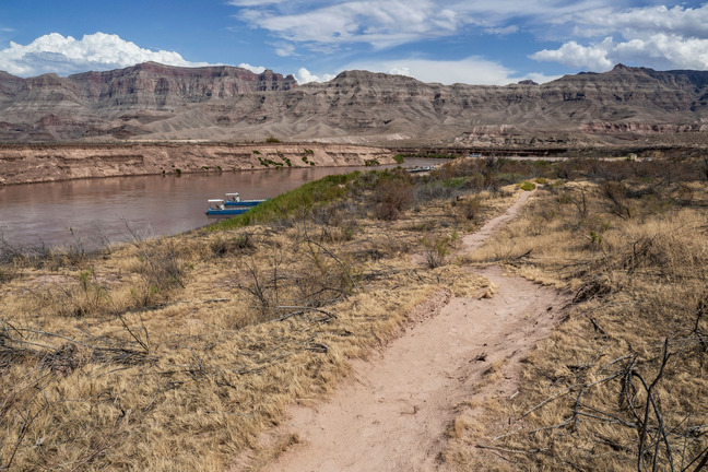 Winding sandy trail on right, river to left, mountains and cloudy sky in background